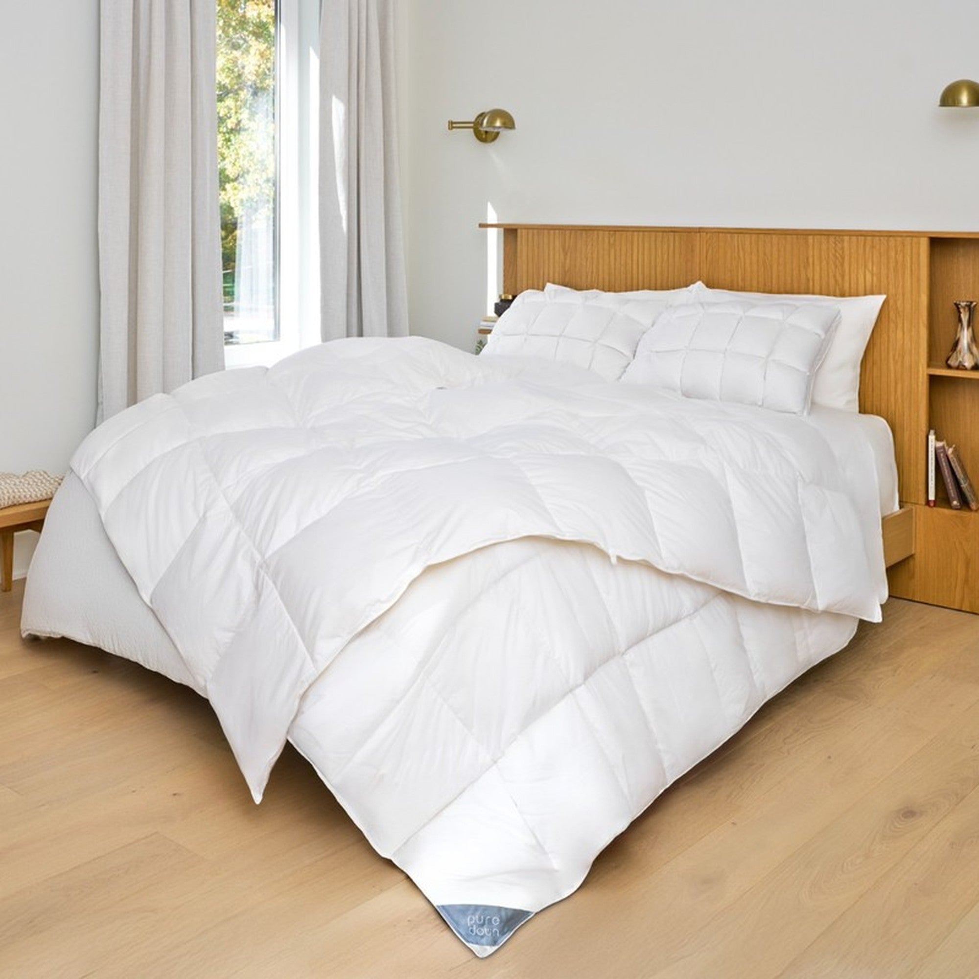 White comforter on a bed in a bedroom with wooden flooring and a window.