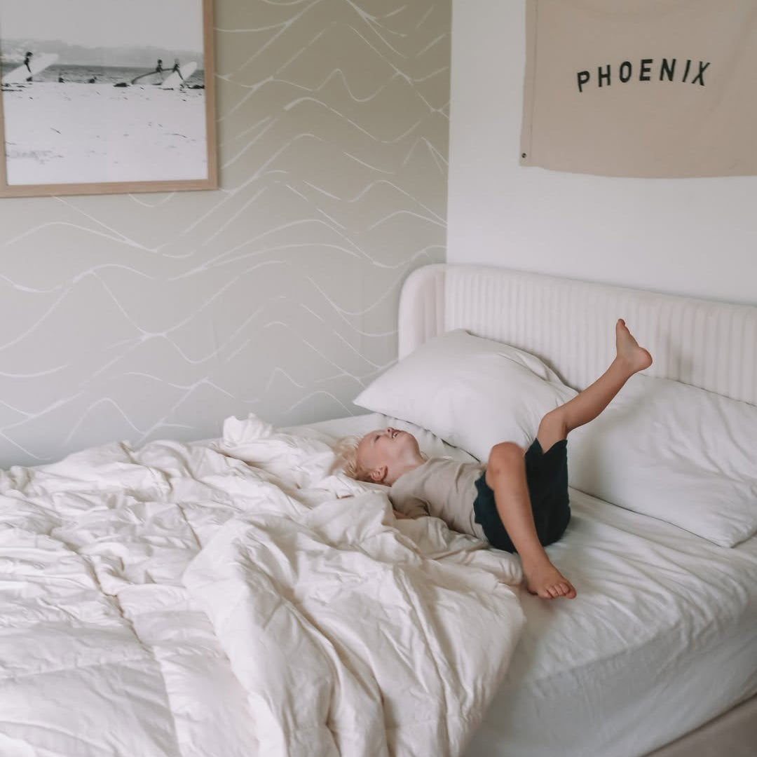 A child lies playfully on a rumpled white bed in a minimal, light-filled bedroom with wall art.
