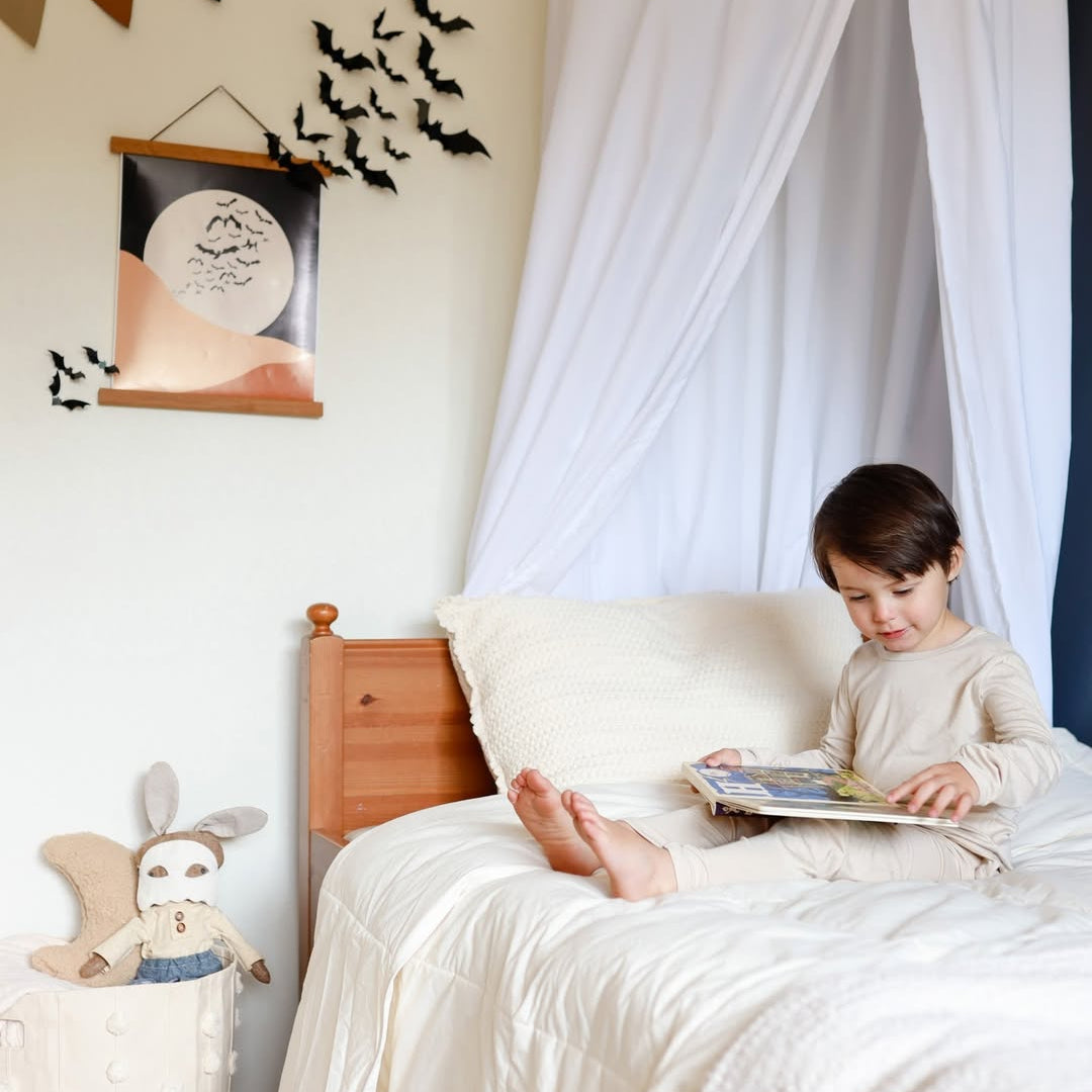 Young child reads on a bed with cream bedding, bat wall decor, and a stuffed rabbit toy nearby.