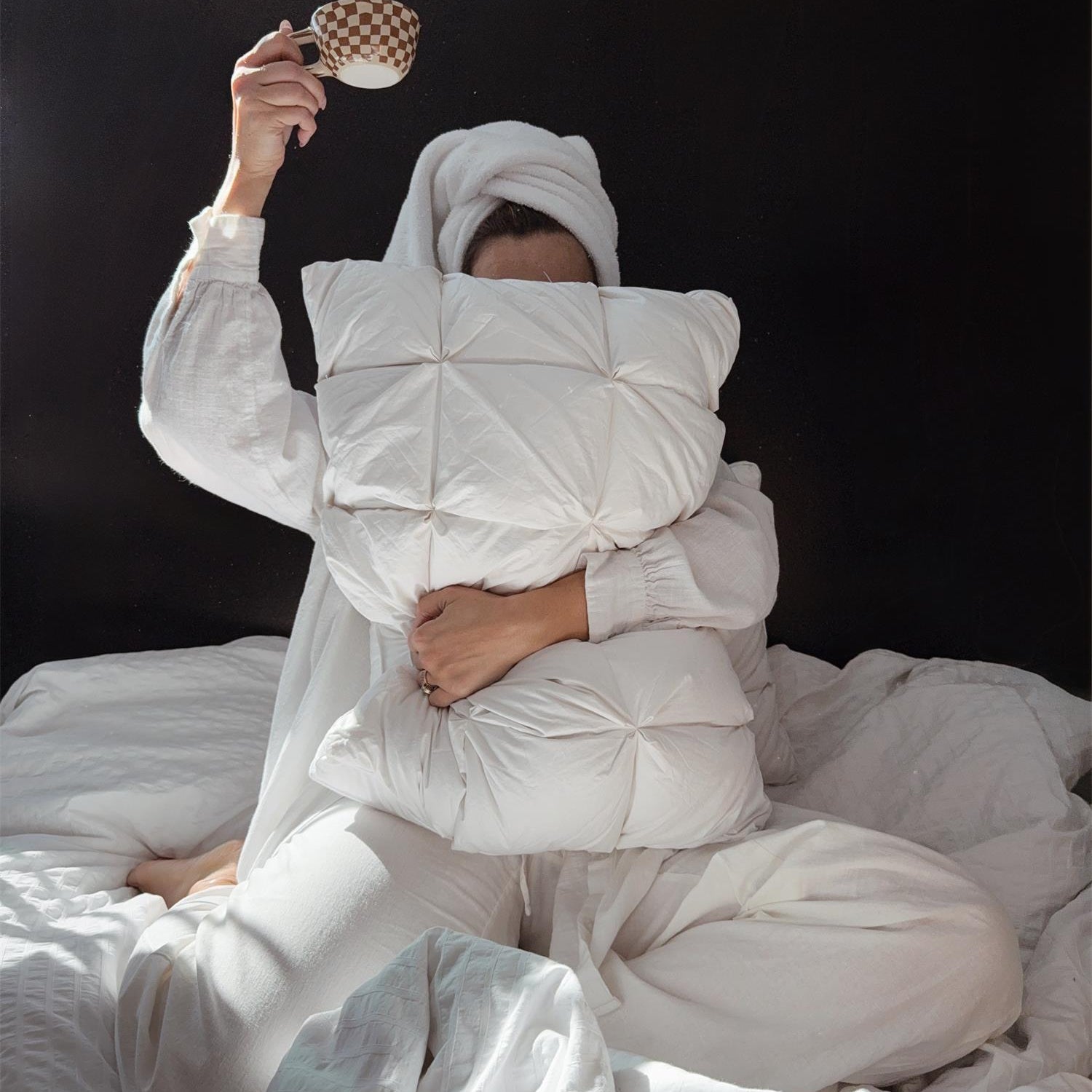 A person in white pajamas and towel holds a pillow and a teacup while sitting on a bed against a dark background.