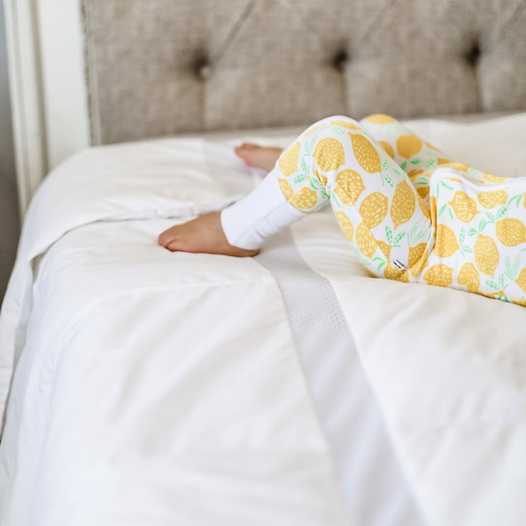 Child in pineapple-print pajamas lying on a white bed, only legs and feet visible.