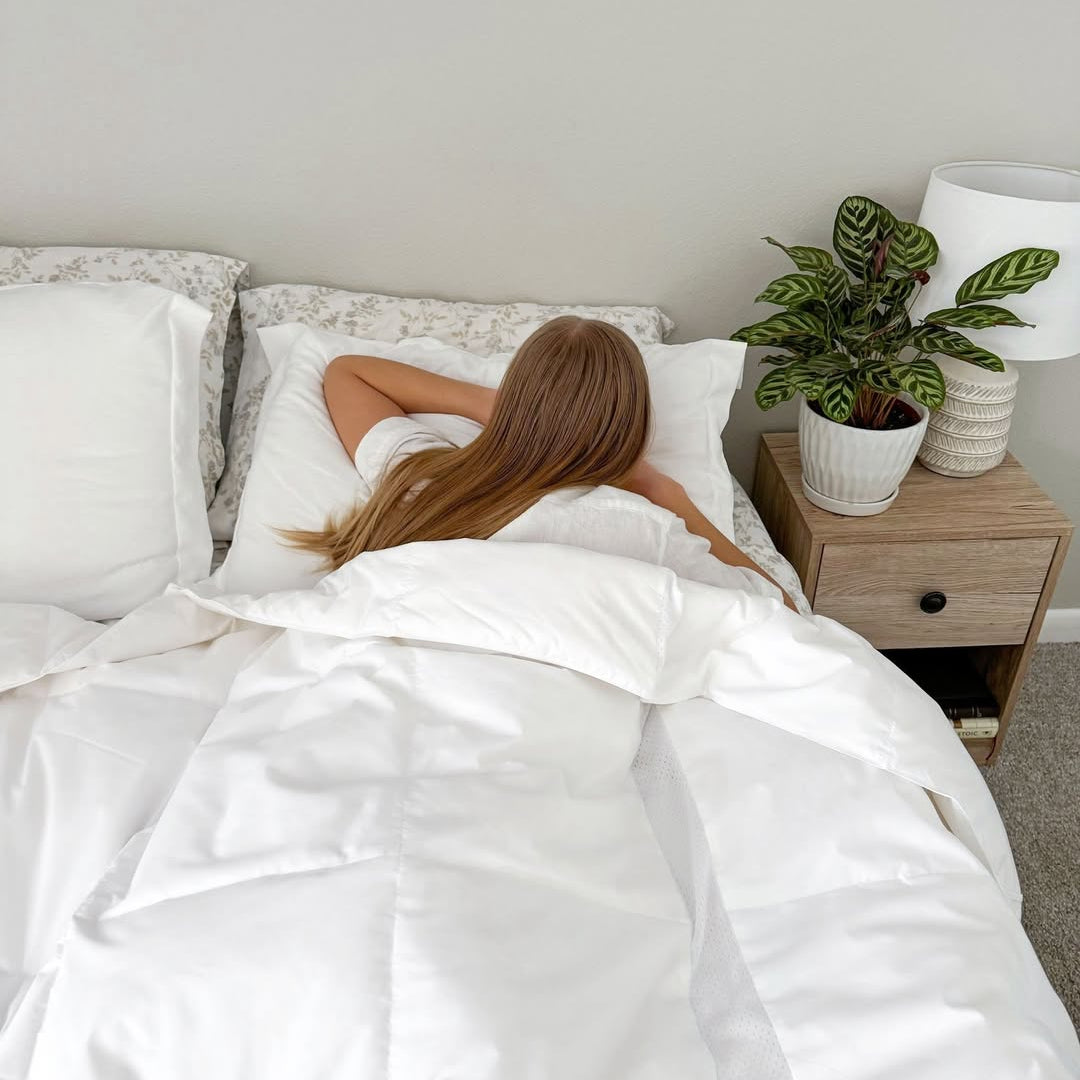 A woman with long hair lies face down in bed under a white comforter beside a nightstand with a potted plant.