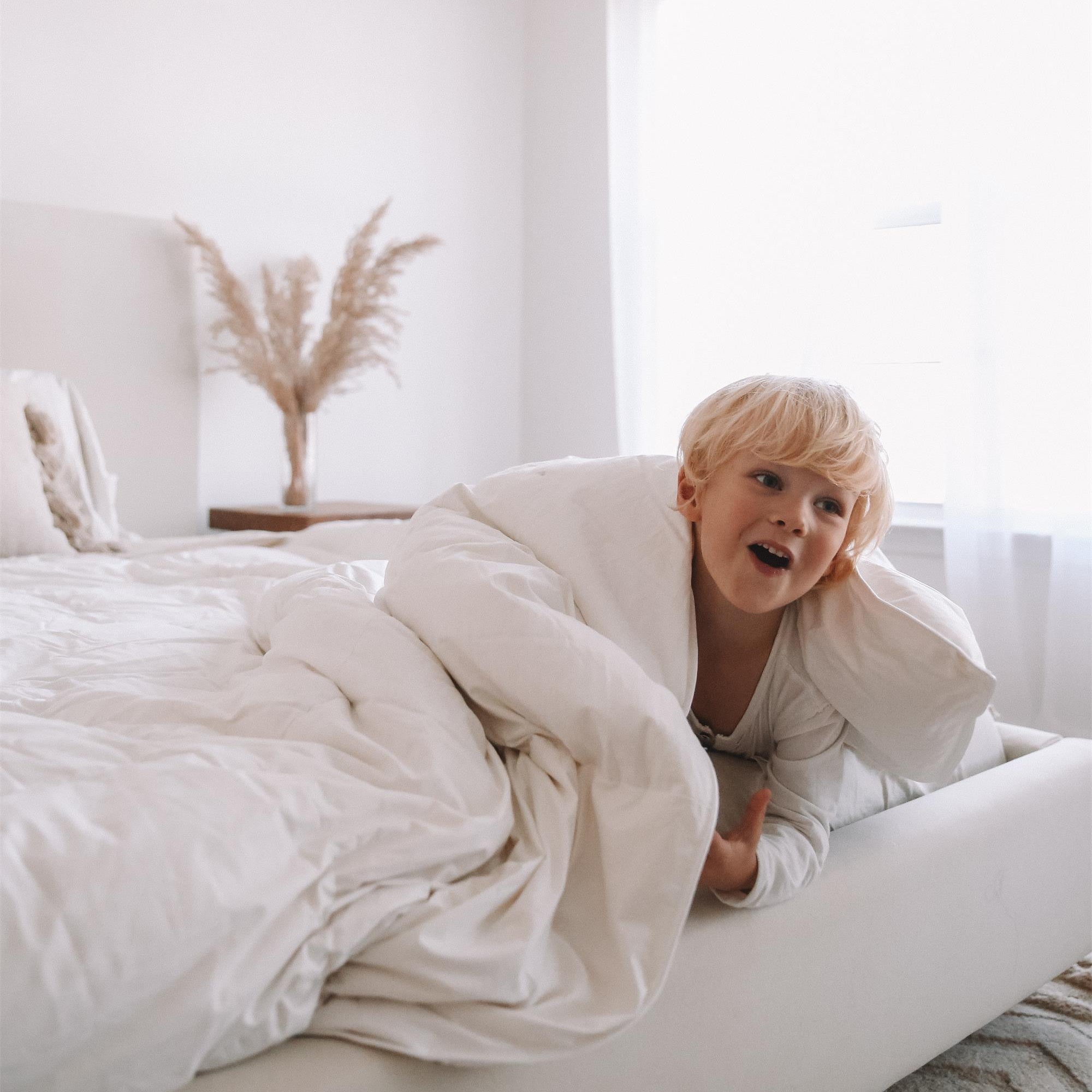 A young child wrapped in a white comforter smiles while lying on a bed in a bright, cozy bedroom.