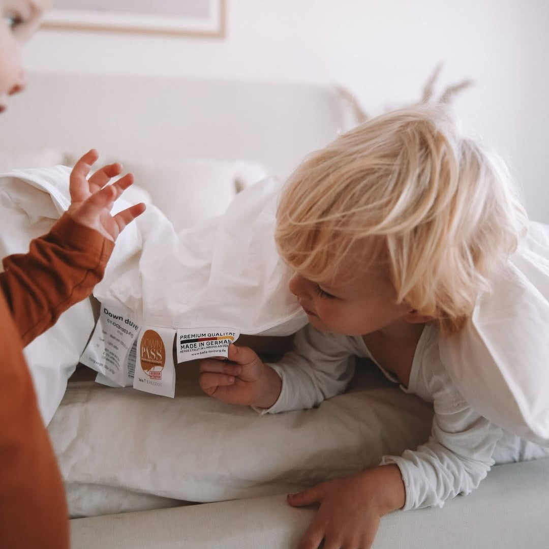 Two young children look at tags on a white comforter while sitting on a bed in a bright room.