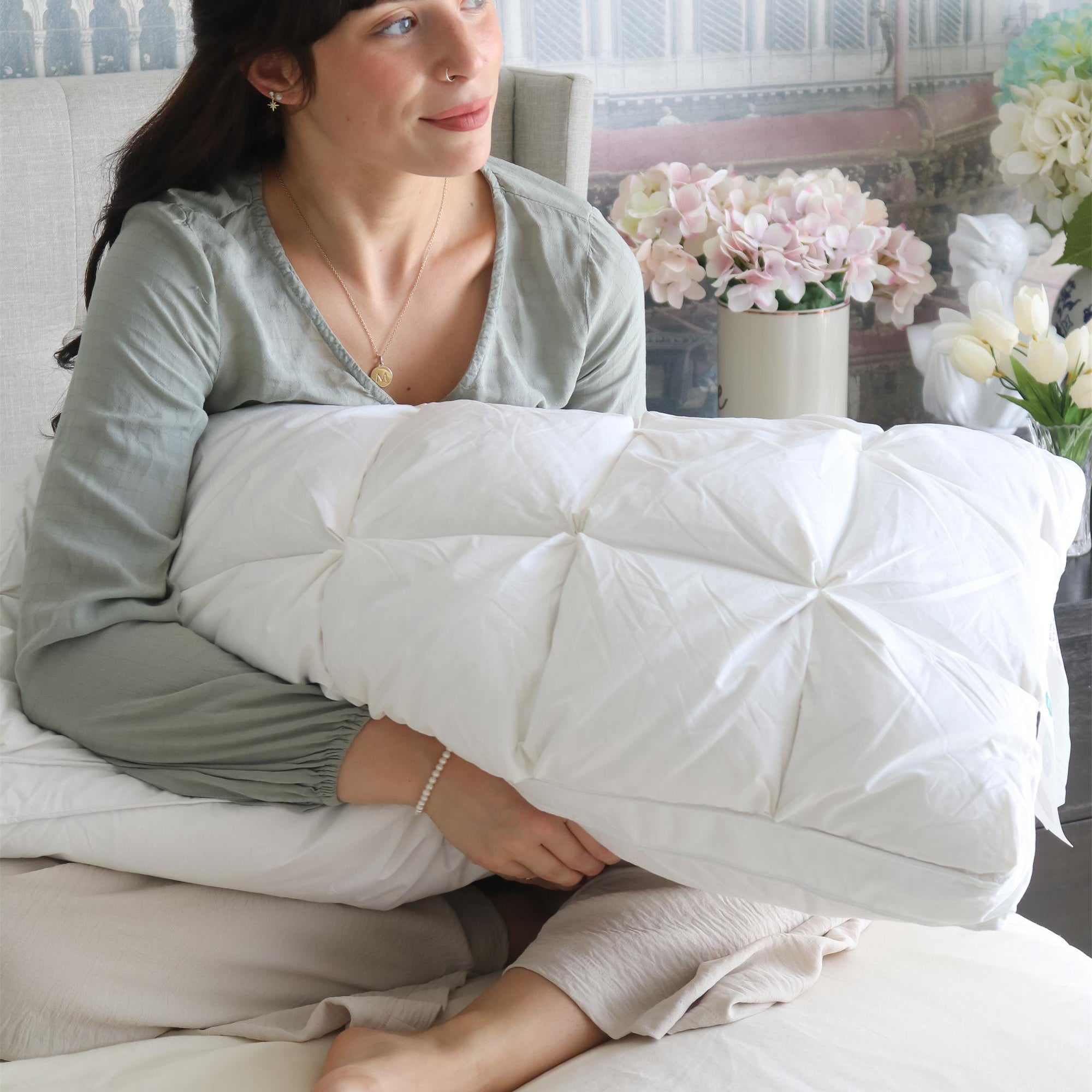 Woman sitting on a bed, hugging a white pillow, looking to the side, with flowers in the background.