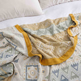 The Boho Cotton Throw Blanket, featuring a patterned jacquard design and yellow trim, is draped over a white bed with pillows in the background.