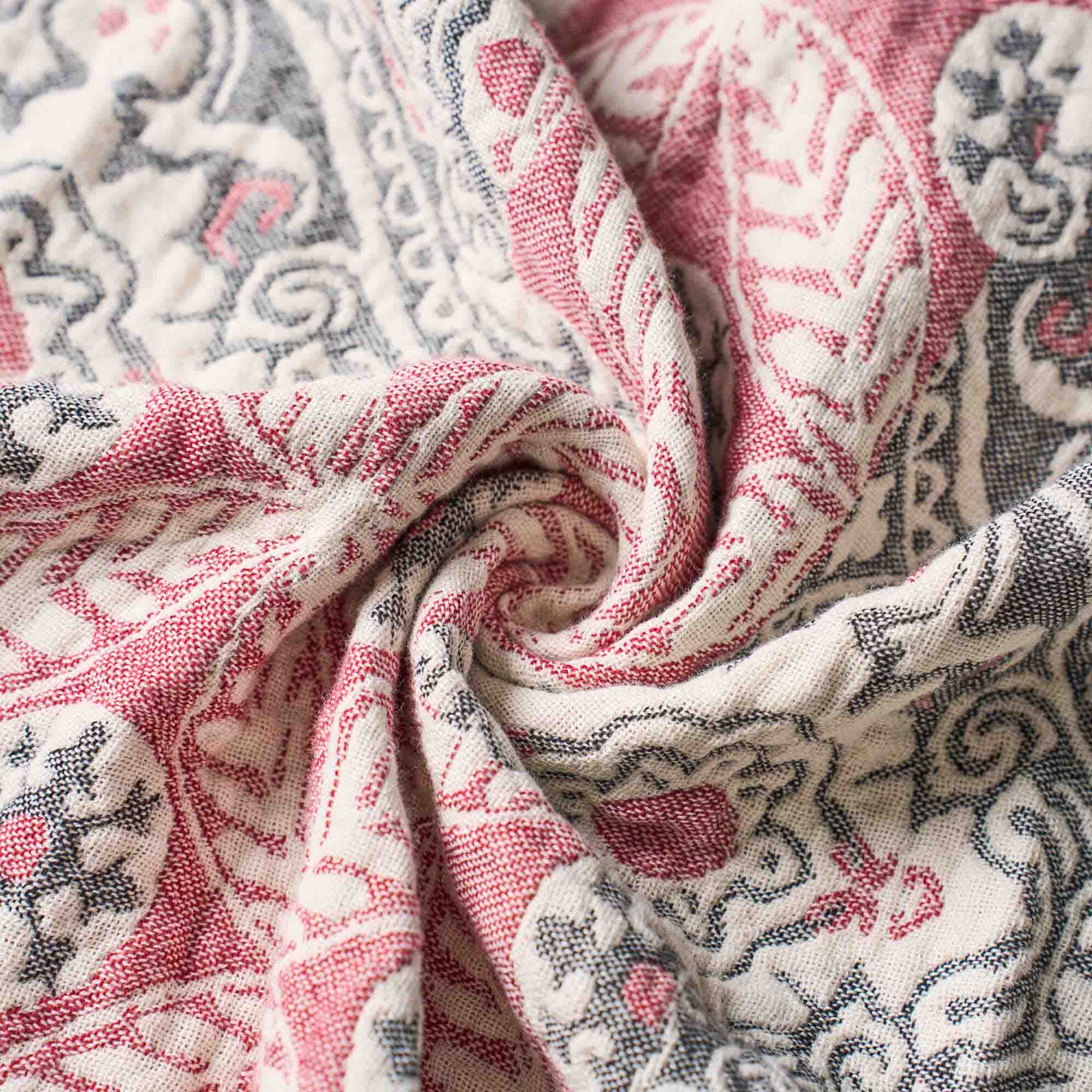 Close-up of textured fabric with red and gray intricate patterns, loosely gathered in the center—the Boho Cotton Throw Blanket featuring a rich, multi-color jacquard design.