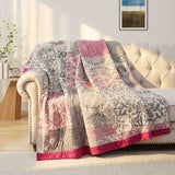 The Boho Cotton Throw Blanket, featuring pink, gray, and white tones in a patterned jacquard design, is draped over a beige tufted sofa in a sunlit room.