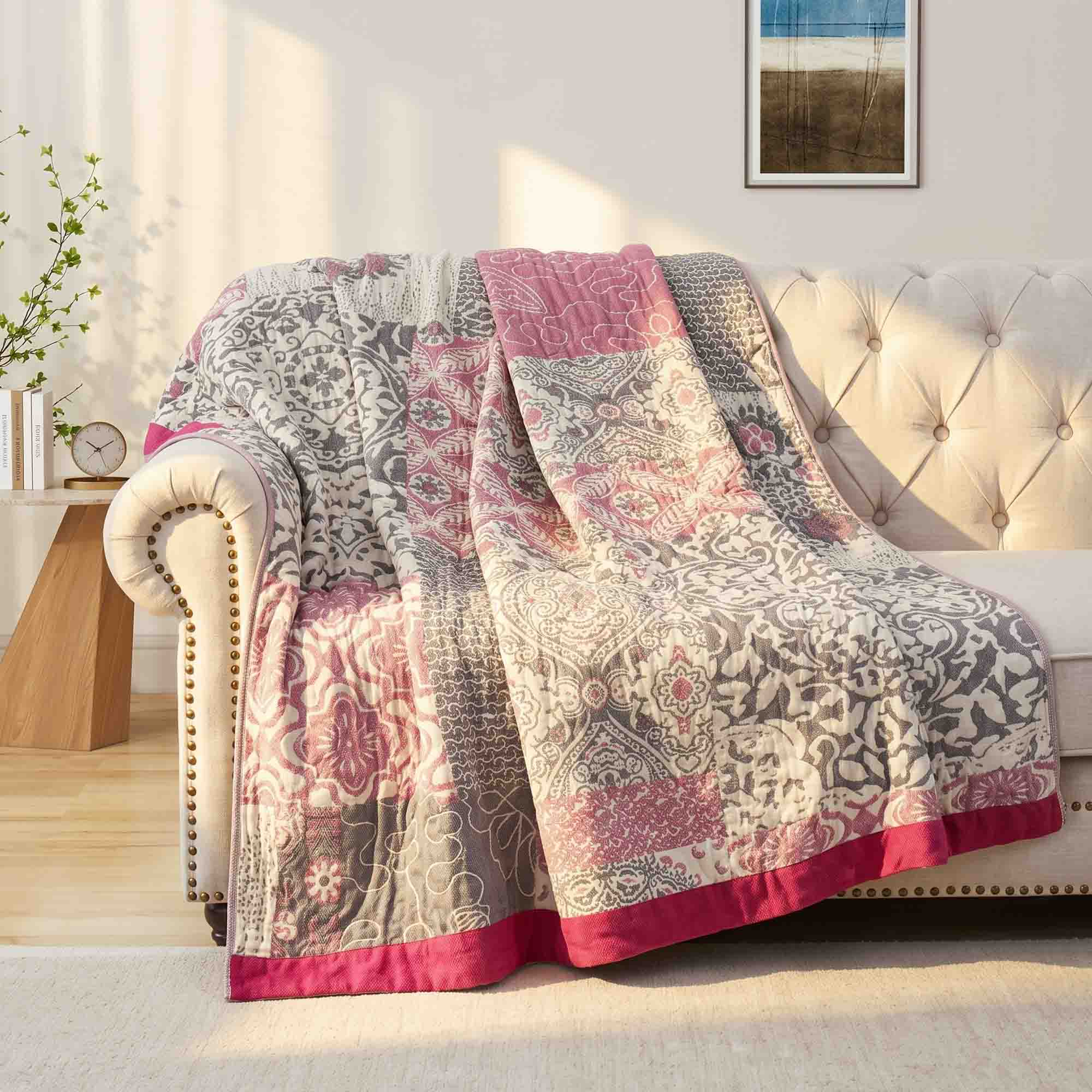 The Boho Cotton Throw Blanket, featuring pink, gray, and white tones in a patterned jacquard design, is draped over a beige tufted sofa in a sunlit room.