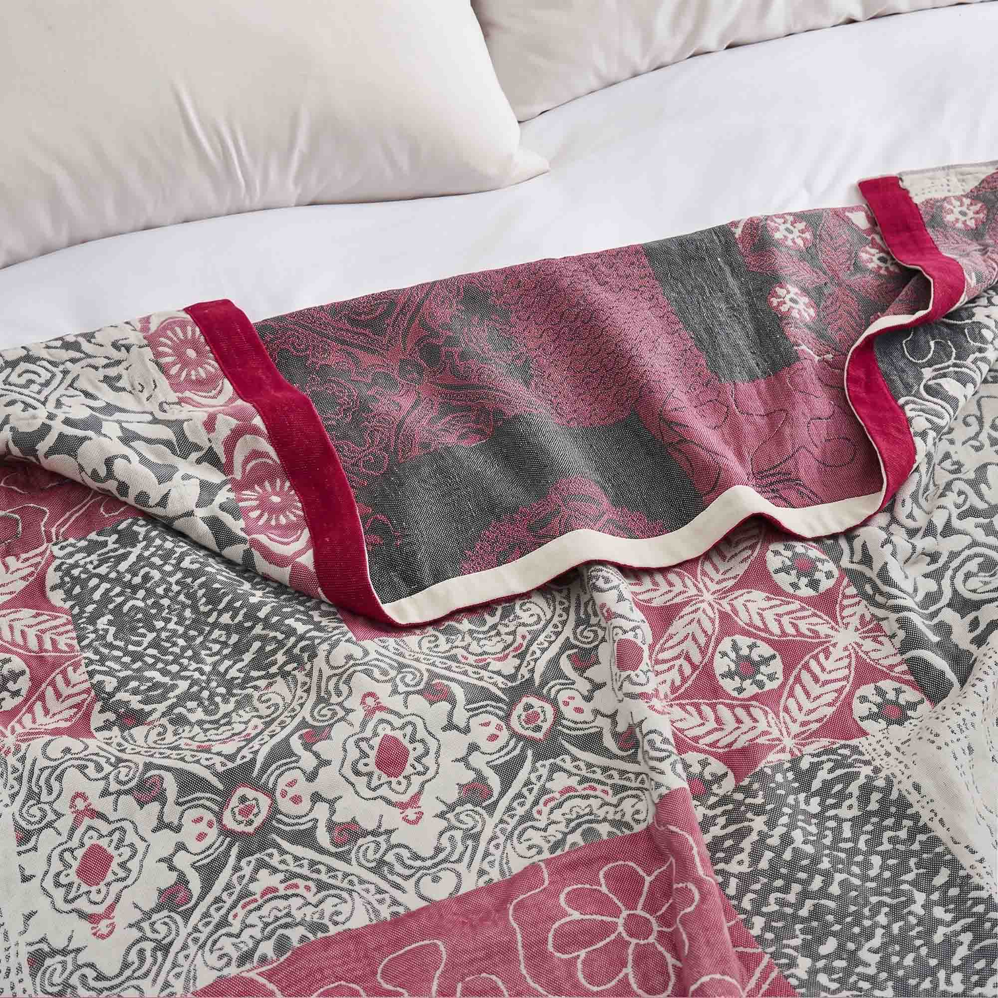 The Boho Cotton Throw Blanket, featuring a red, gray, and white floral jacquard pattern, is spread across a white bed with pillows.