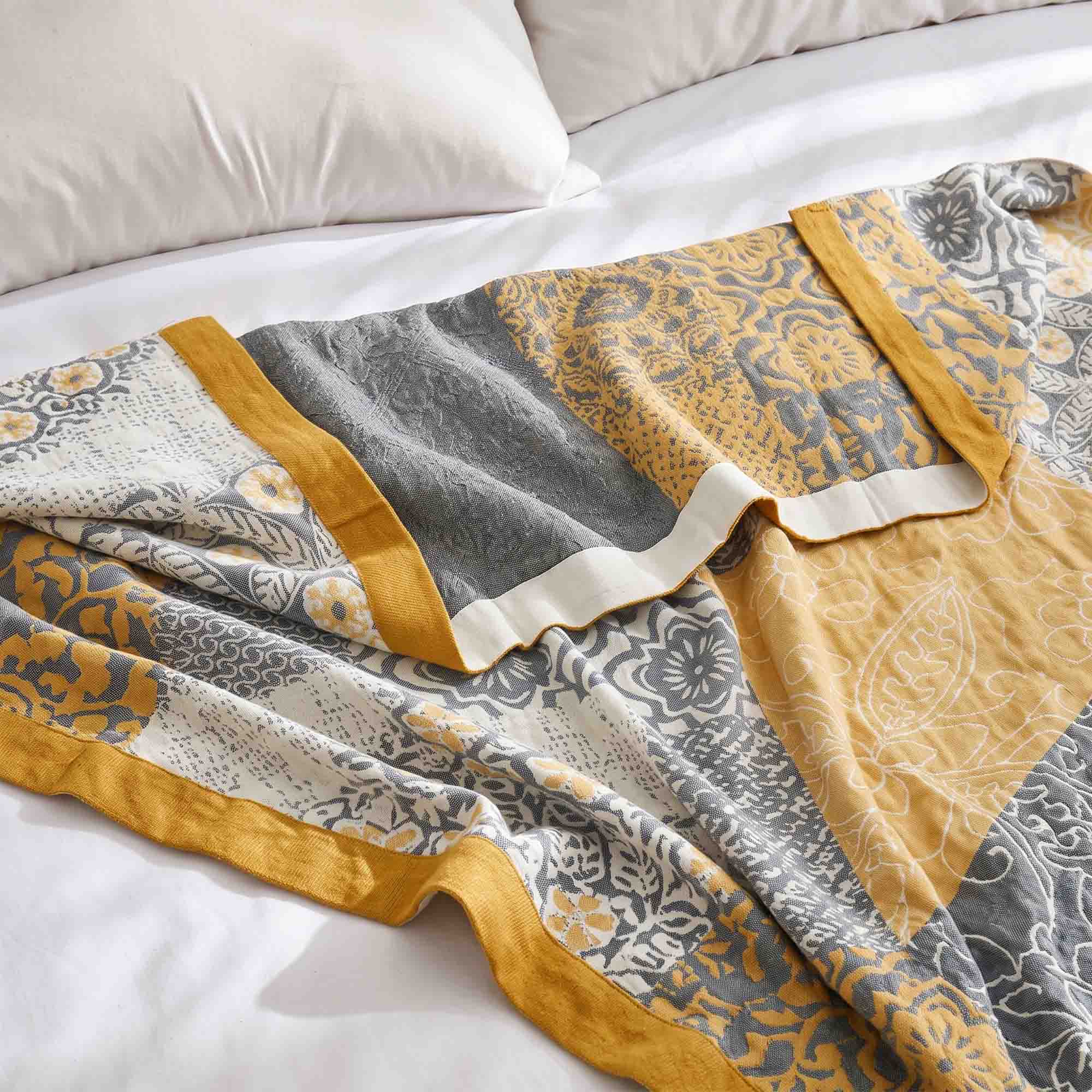 The Boho Cotton Throw Blanket, featuring a patterned yellow, gray, and white jacquard design, is draped over a white bed adorned with pillows.