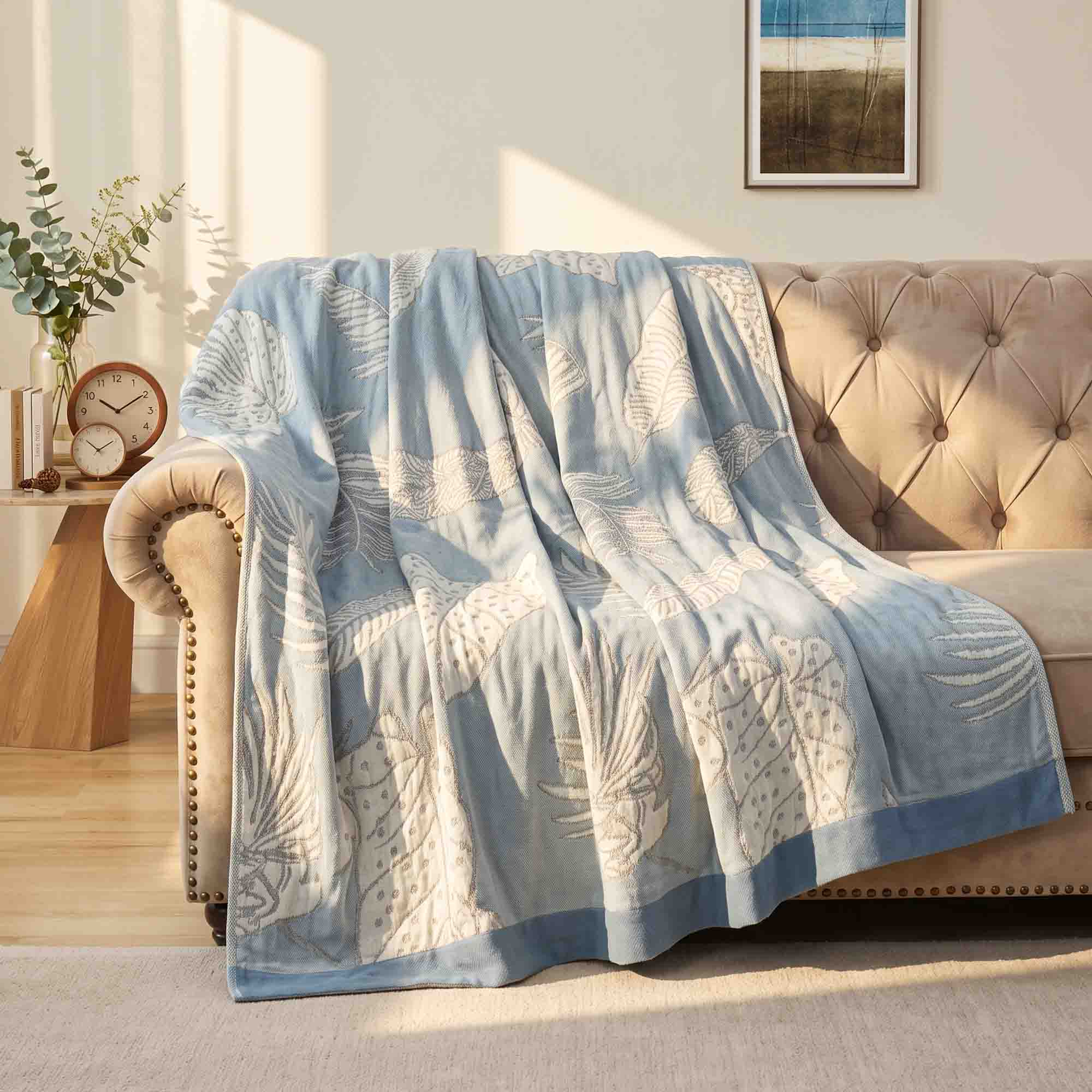 A Boho Cotton Throw Blanket featuring blue and white patterns is draped over a beige tufted sofa in a sunlit living room.