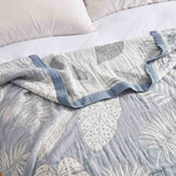 A Boho Cotton Throw Blanket with a blue and white leaf pattern lies on a bed with white pillows in the background.