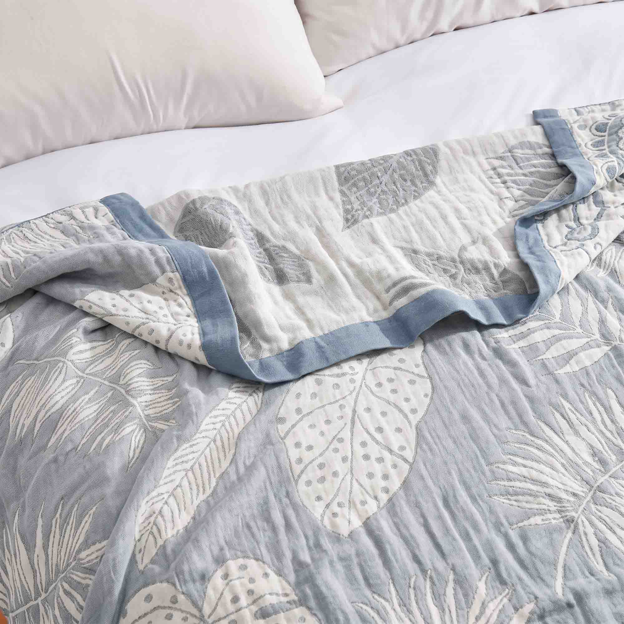 A Boho Cotton Throw Blanket with a blue and white leaf pattern lies on a bed with white pillows in the background.