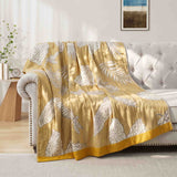 The Boho Cotton Throw Blanket, featuring a yellow and white leaf pattern in cotton jacquard, is draped over a light couch in a sunny, modern living room.