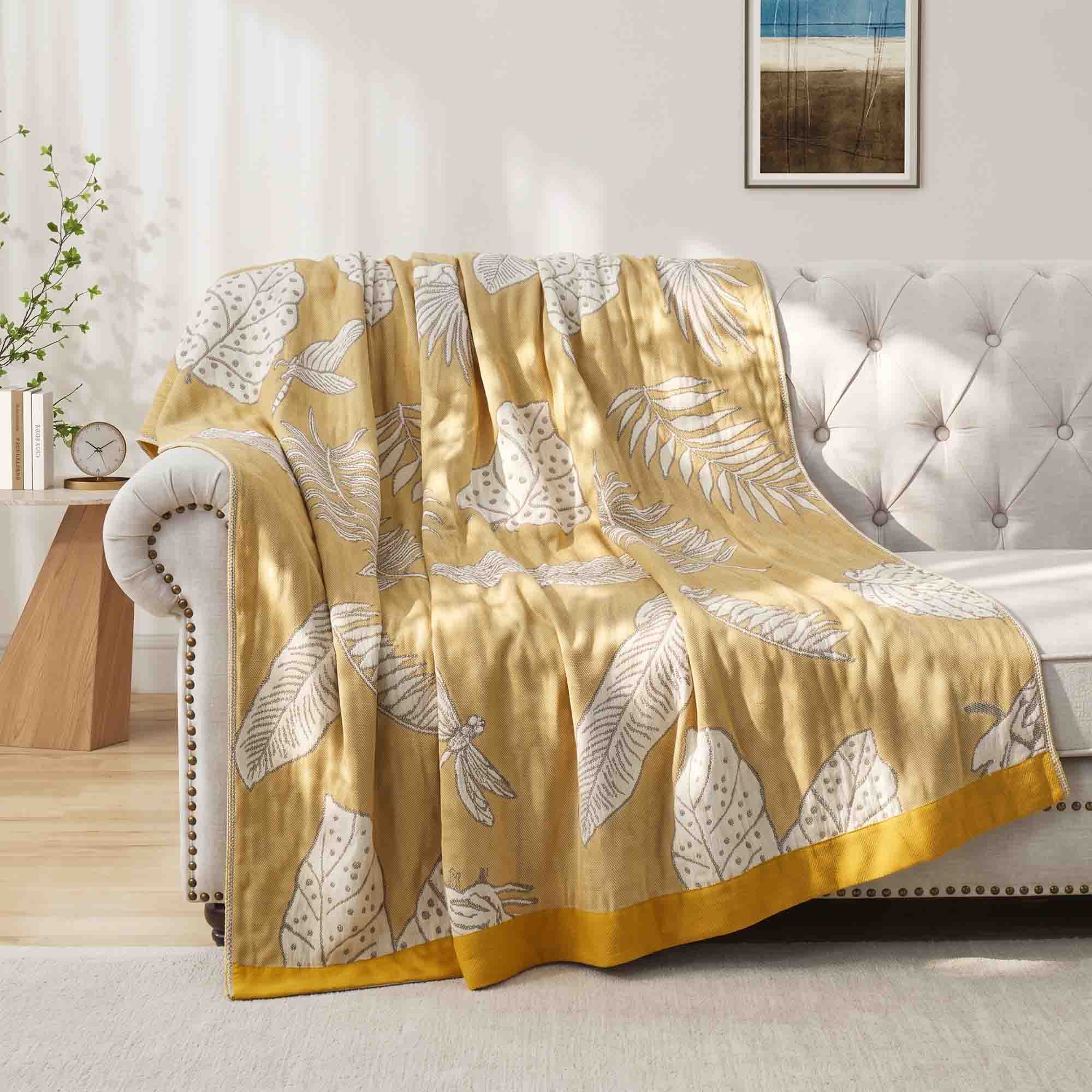 The Boho Cotton Throw Blanket, featuring a yellow and white leaf pattern in cotton jacquard, is draped over a light couch in a sunny, modern living room.