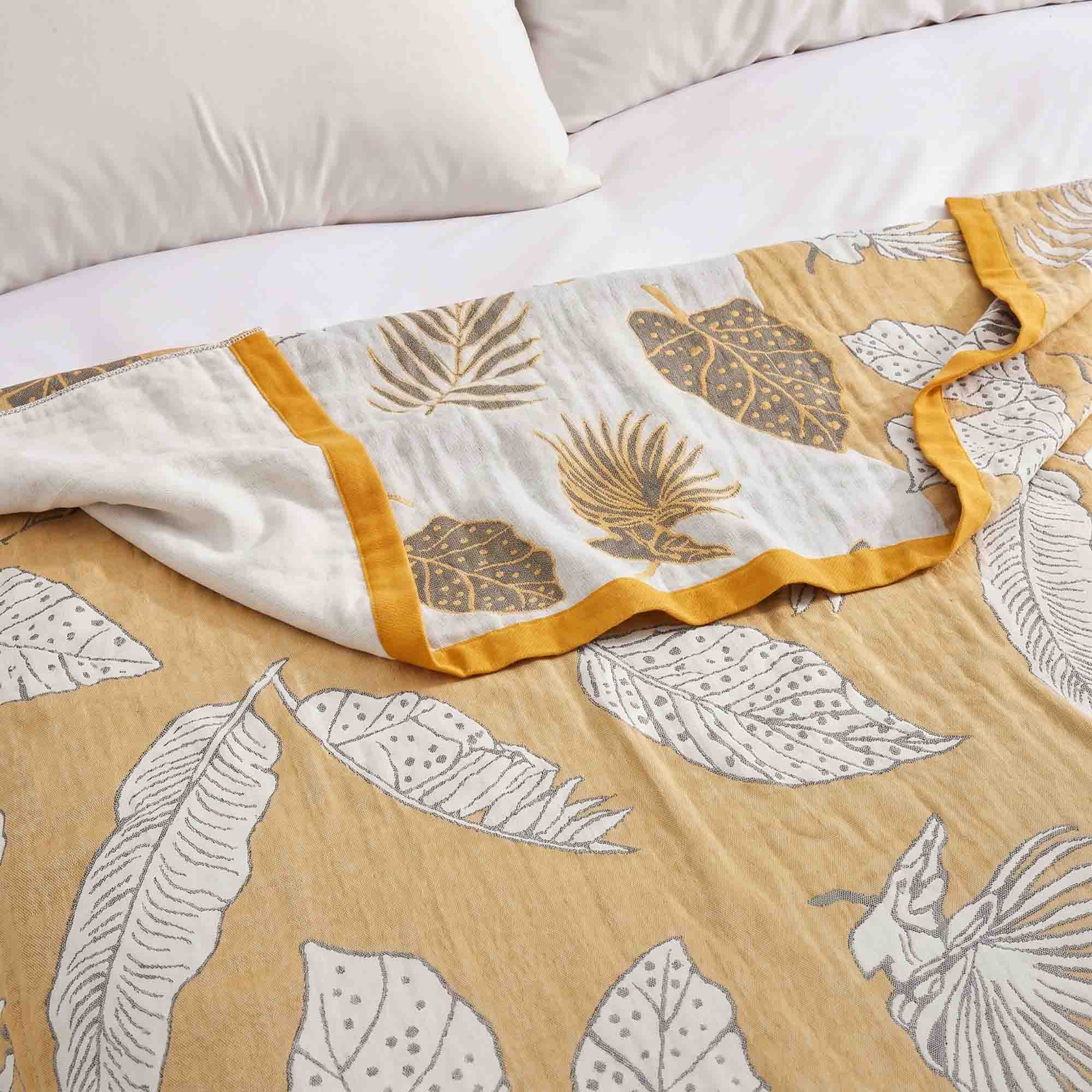 A Boho Cotton Throw Blanket with yellow and white leaf jacquard patterns is spread on a bed with white pillows.