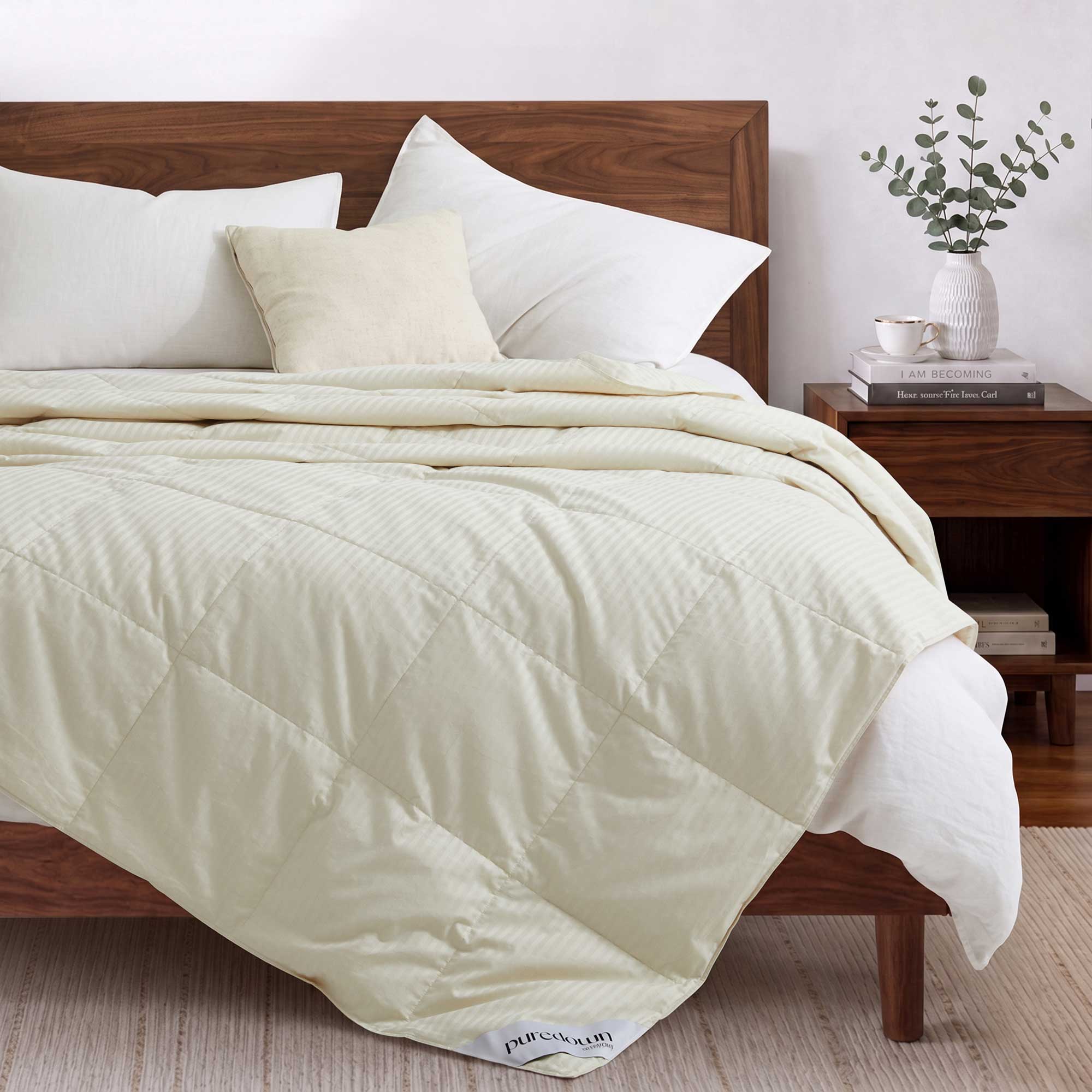 A neatly made bed features the Woven Stripe Down Blanket in cream, complemented by white pillows, a wooden headboard, and a nightstand adorned with books and a vase.