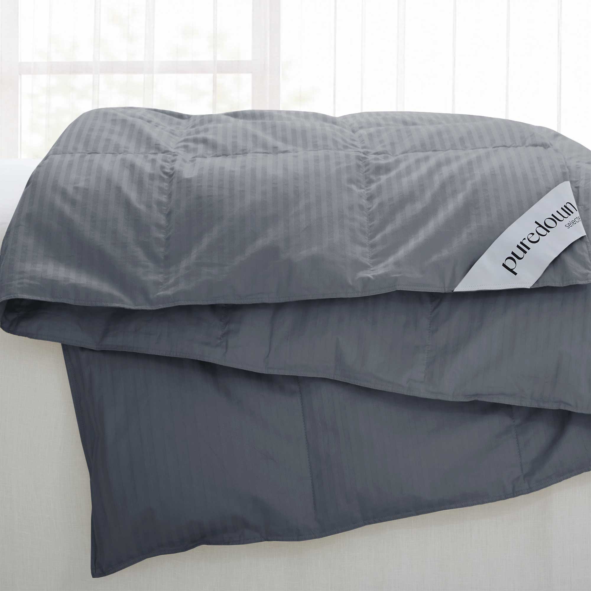 A Woven Stripe Down Blanket in dark gray with a puredown label is draped over a white couch near a window.