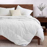 A neatly made bed featuring the Woven Stripe Down Blanket with white bedding, a wooden headboard, pillows, and a vase with leafy branches on the nightstand.