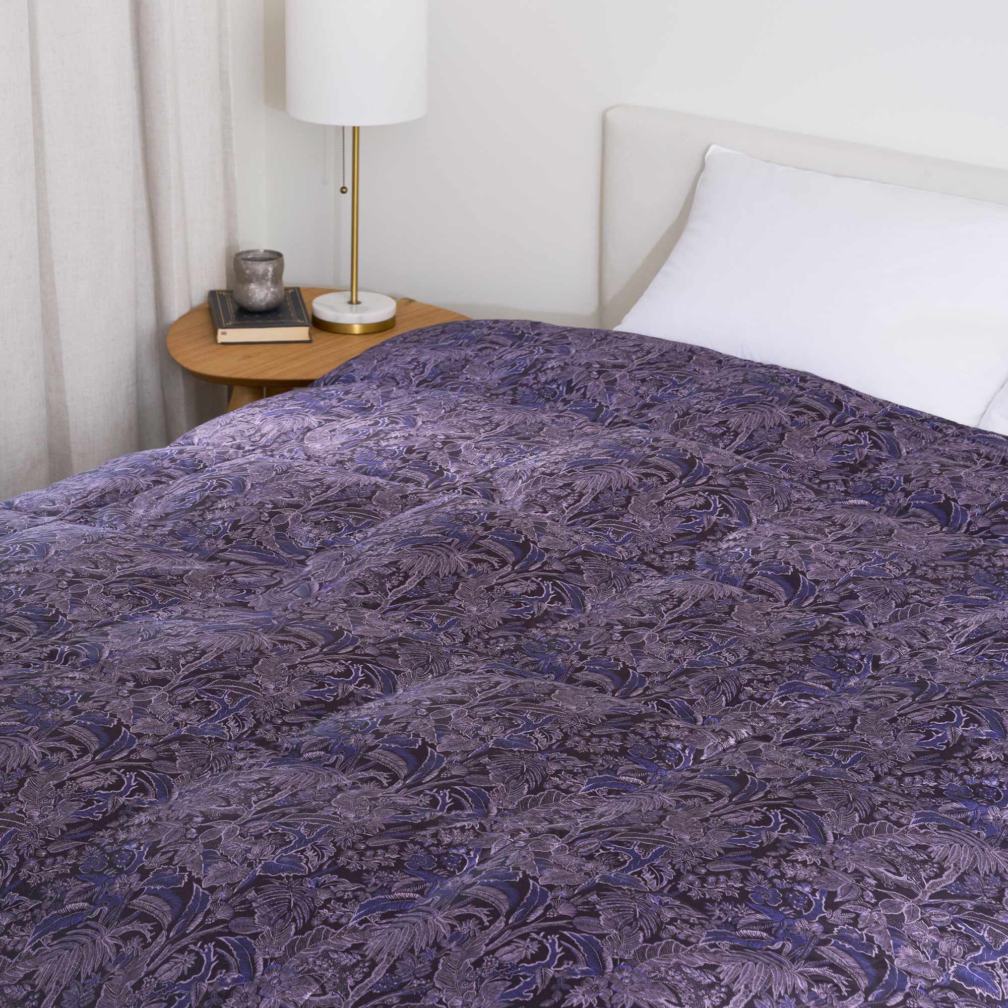 A bed with a purple patterned comforter, white pillow, and a wooden nightstand with a lamp and decor.