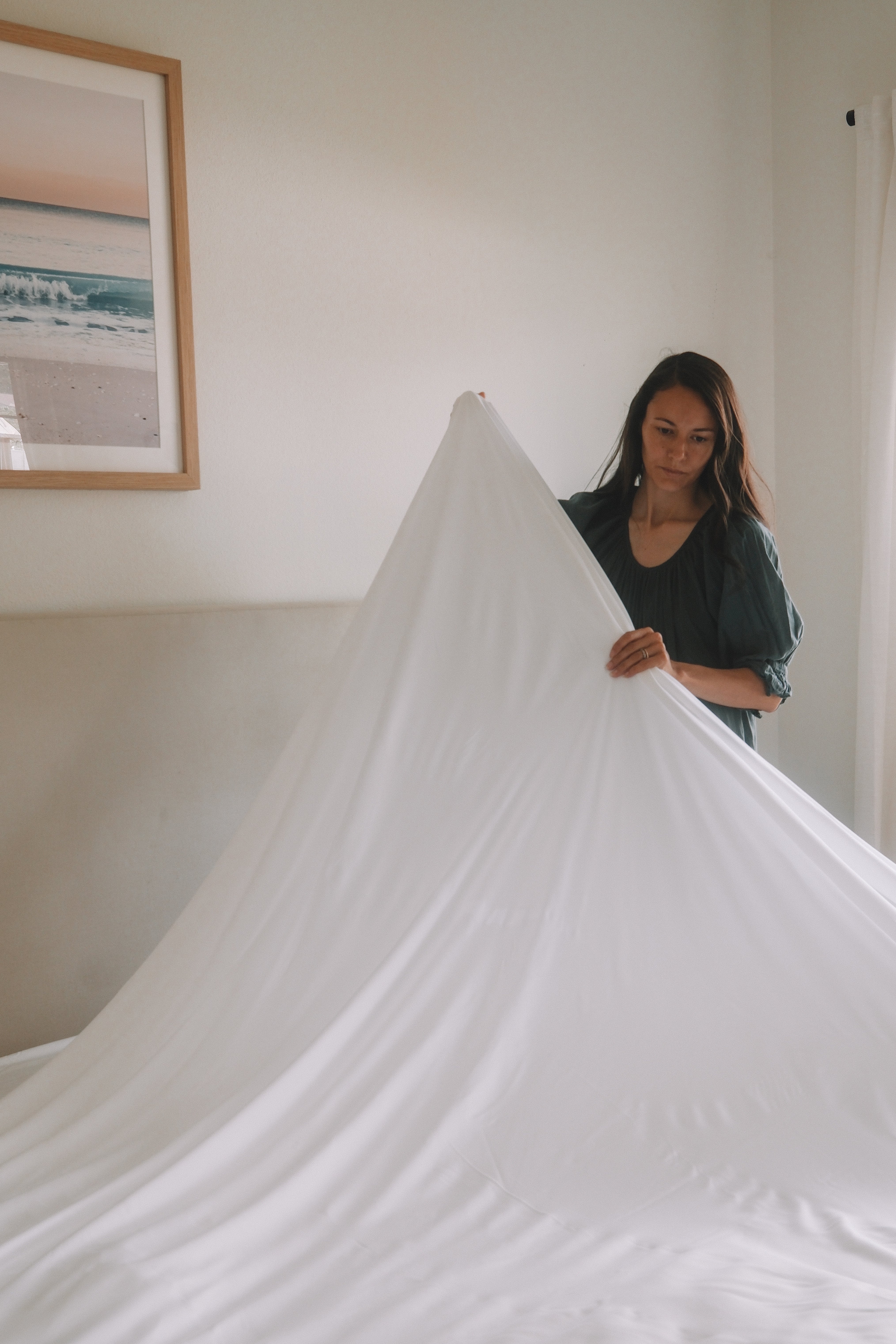 Woman making bed by stretching a white organic bamboo bedsheet over a mattress in a minimalist bedroom with neutral tones and framed beach artwork on the wall.