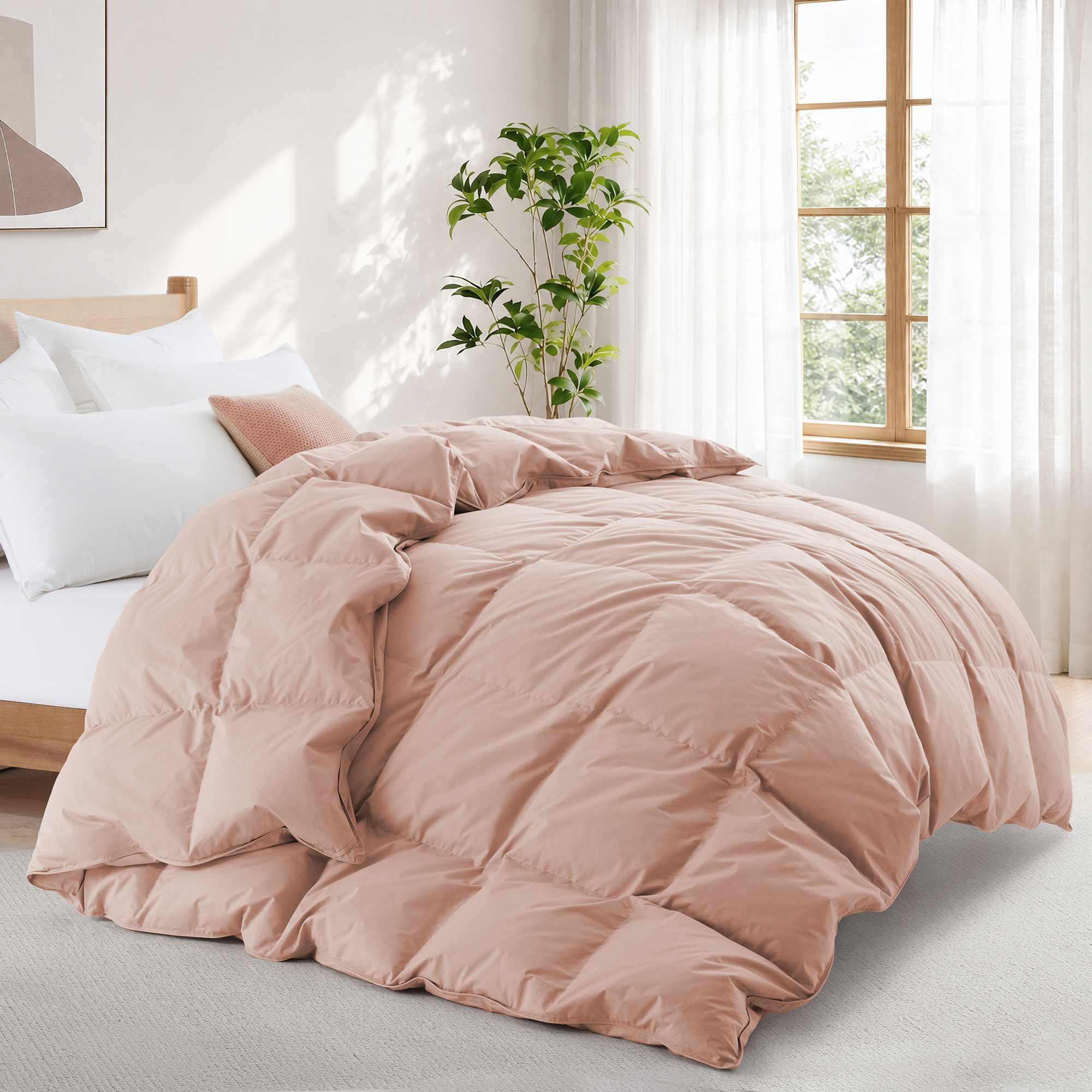 A neatly made bed features the Premium Goose Feather Comforter in light pink, set in a bright, airy bedroom with large windows and a plant.