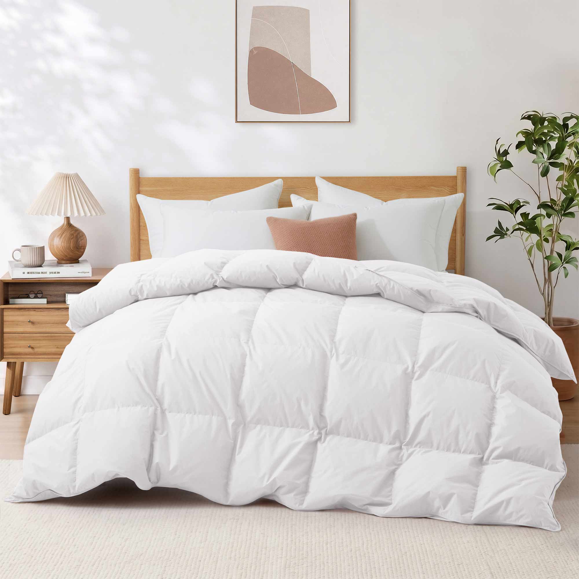 A neatly made bed featuring the Premium Goose Feather Comforter in white bedding, paired with a wood headboard, nightstand, lamp, plant, and abstract wall art.