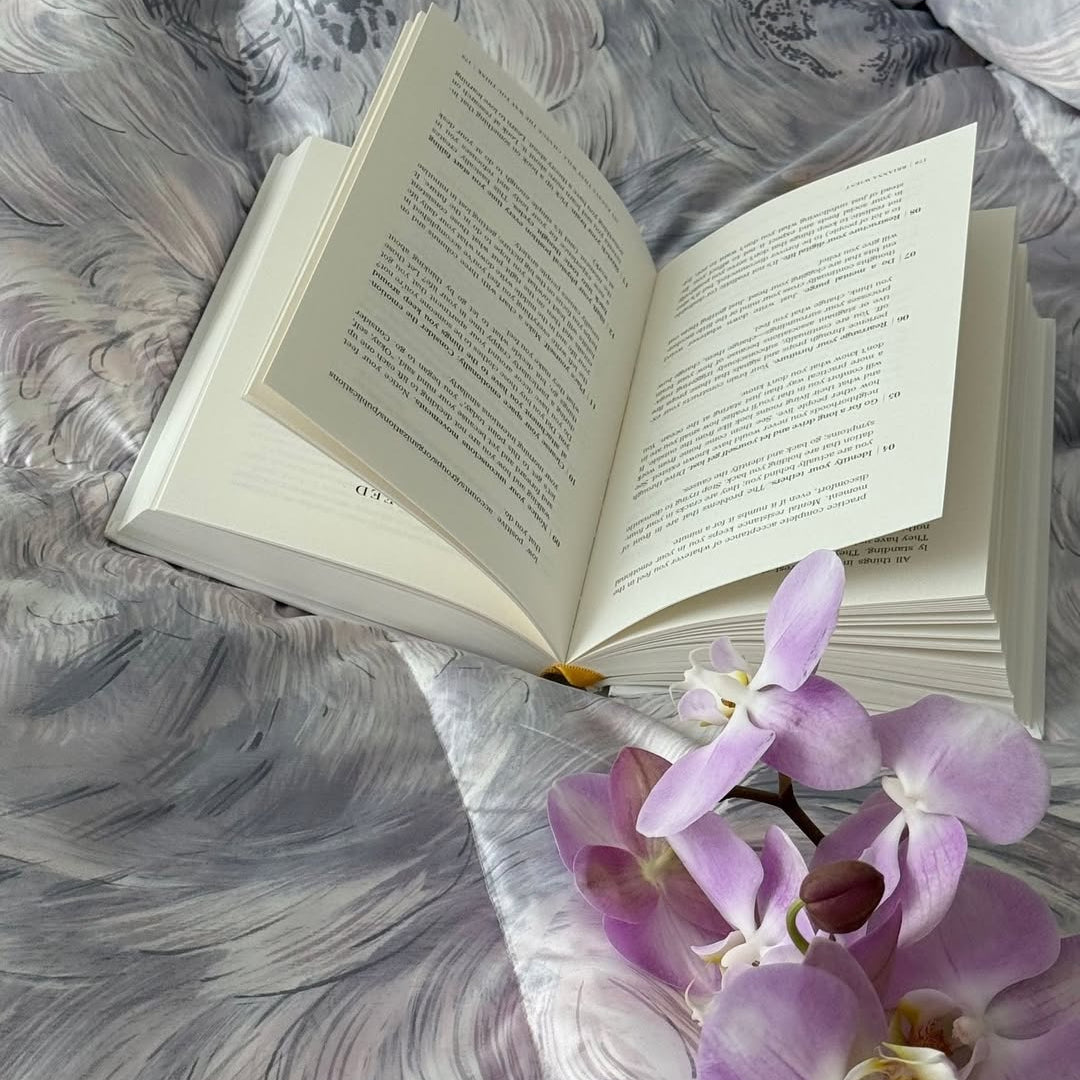 An open book and a pink orchid rest on a gray, patterned blanket.