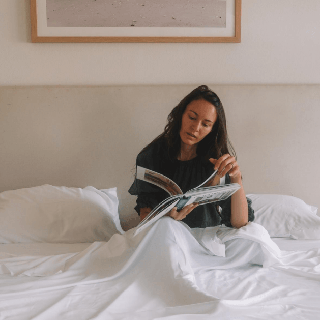 Woman sitting on a bed reading a magazine, with a beach photo hanging on the wall behind her.