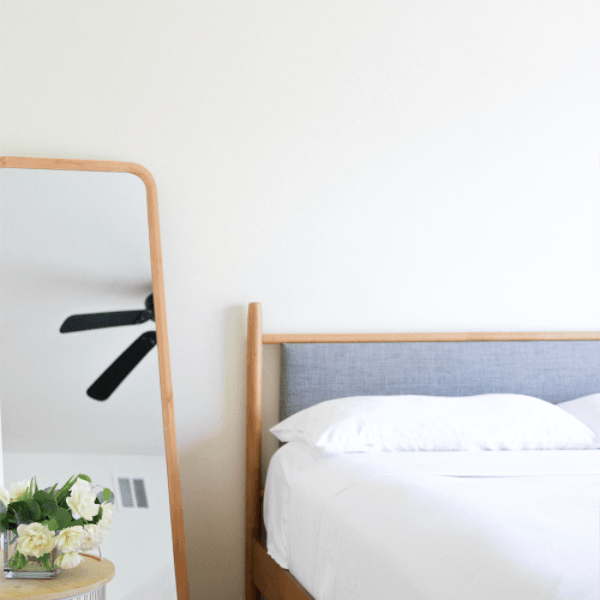 Minimalist bedroom with a bed, leaning mirror reflecting a ceiling fan, and flowers on a side table.