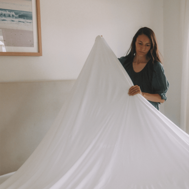 Woman spreading a white sheet over a bed in a softly lit room with a beach photo on the wall.