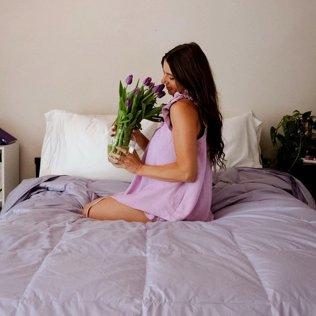 Woman in a lavender nightgown sits on a bed, holding a vase of purple tulips and smiling.