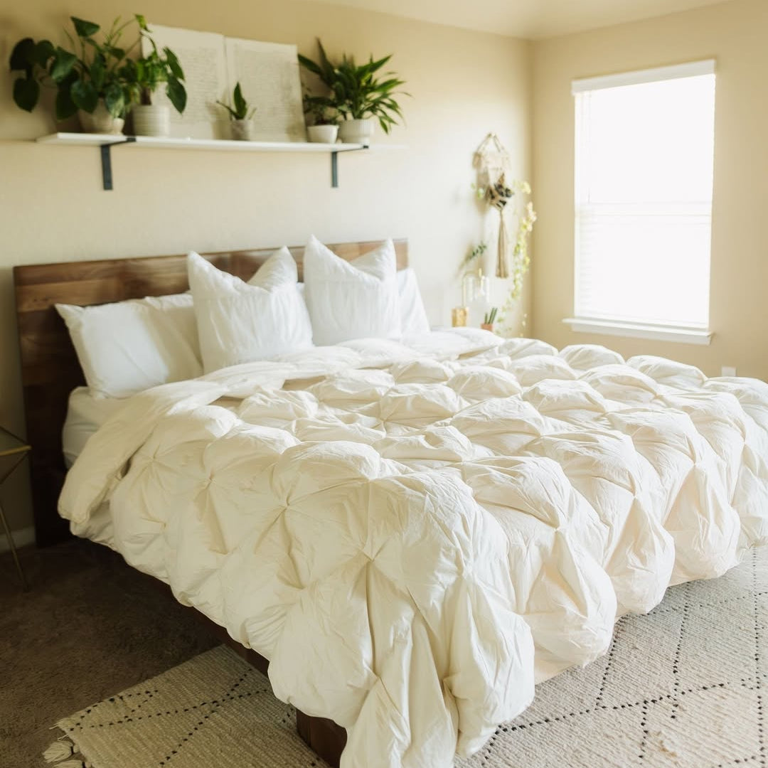 Cozy bedroom with a fluffy white comforter, white pillows, potted plants, and sunlight through a window.