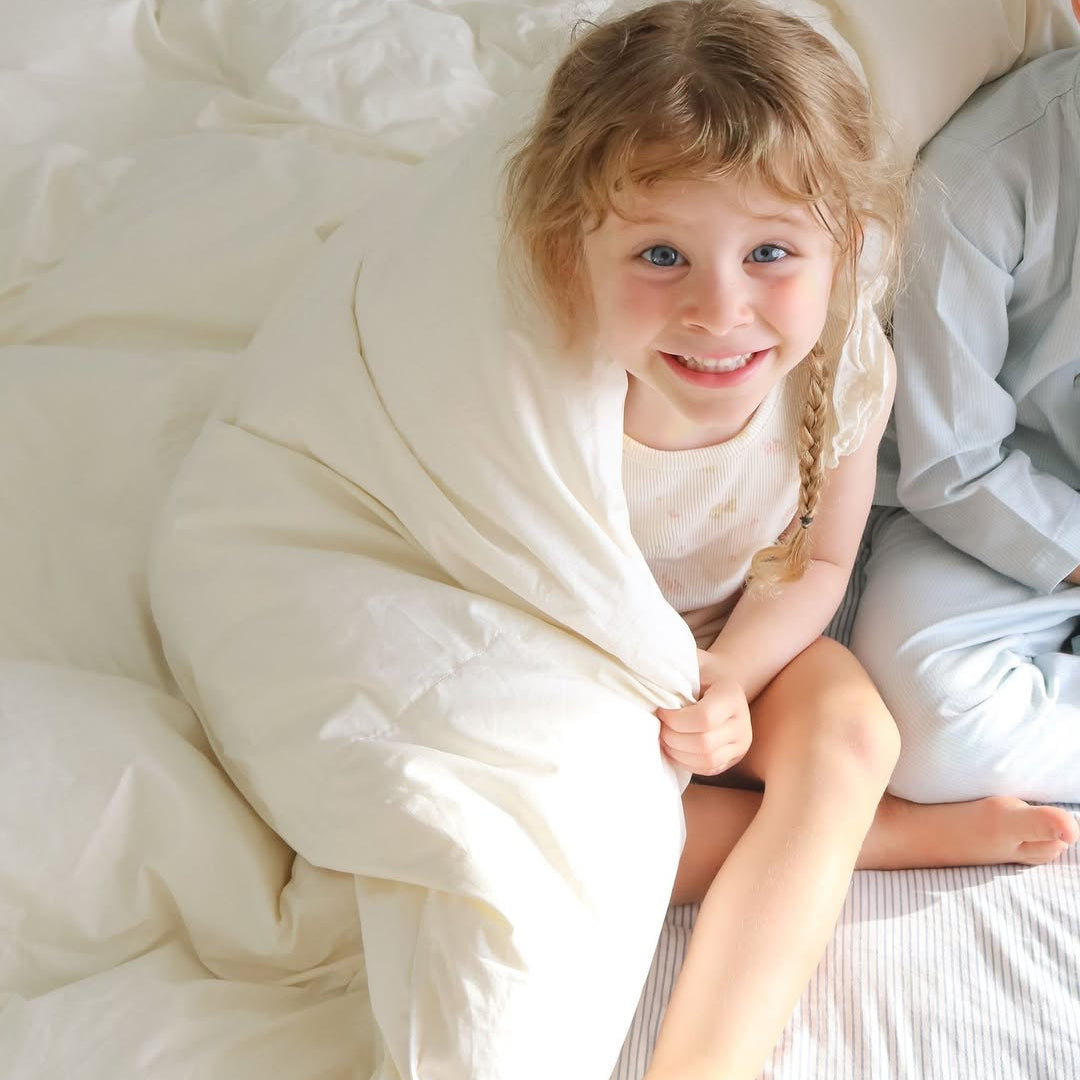 Smiling child wrapped in a white blanket, sitting on a bed with sunlight illuminating their face.