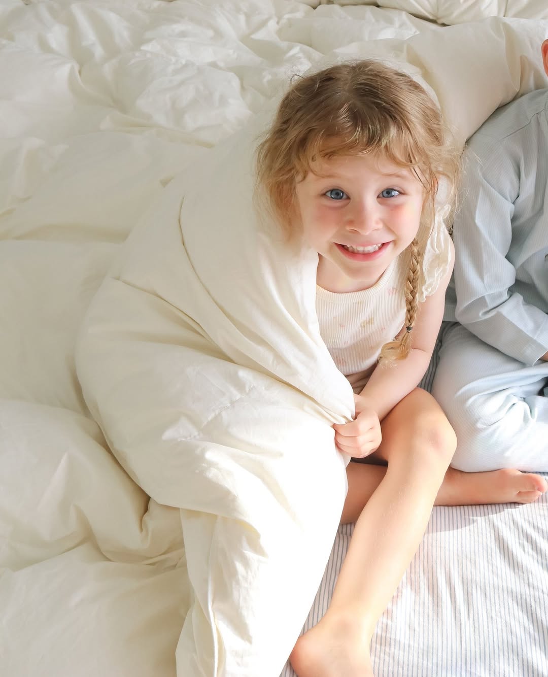 Smiling child wrapped in a white blanket, sitting on a bed with sunlight illuminating their face.