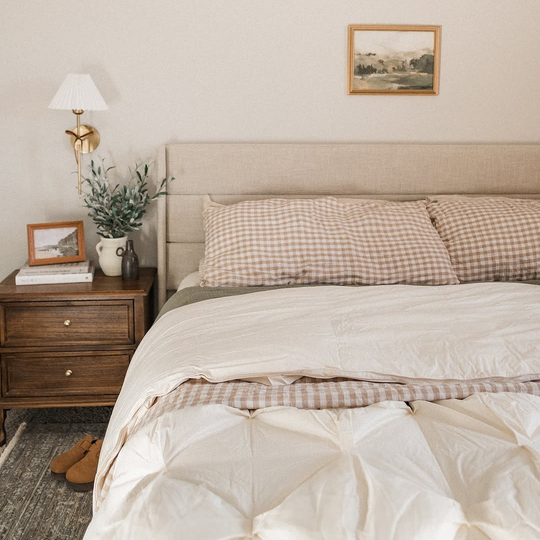 Cozy bedroom with beige bedding, gingham pillows, a wooden nightstand, and framed art on the wall.