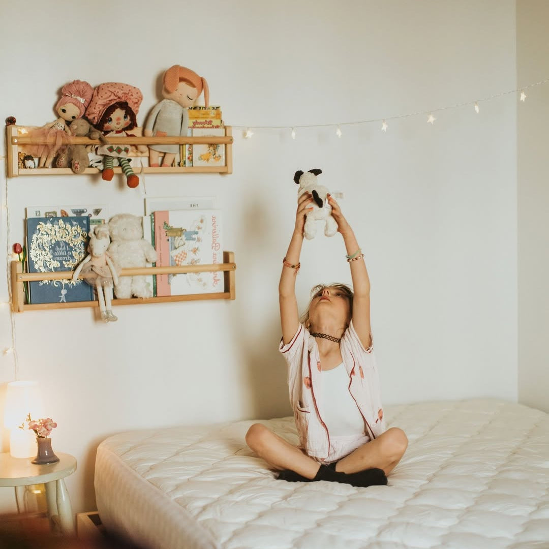 Child sitting on a bed holding a stuffed animal up, with shelves of dolls and books on the wall behind.