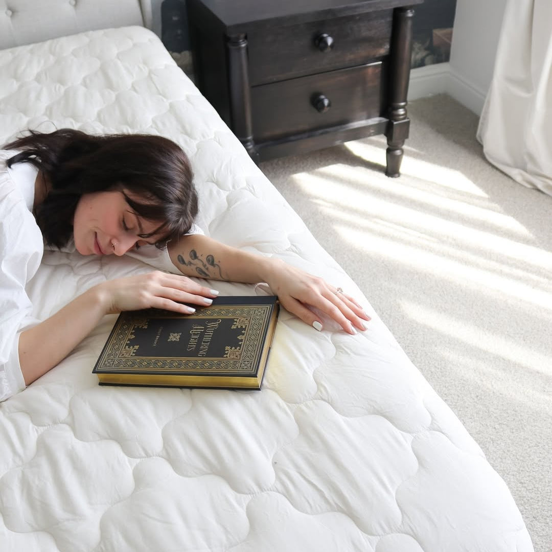 Woman lying on a white mattress, holding a book, with a visible stain on the mattress beside her.